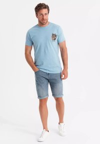 Camiseta de manga corta azul claro con un diseño gráfico en el bolsillo del pecho izquierdo, combinada con pantalones cortos de mezclilla enrollados y zapatillas blancas.