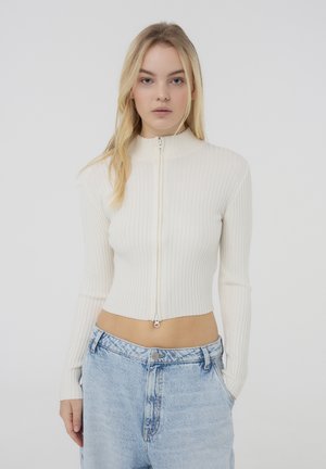 Maglione corto zip-up a coste color crema con colletto alto e maniche lunghe, abbinato a jeans larghi di color blu chiaro.