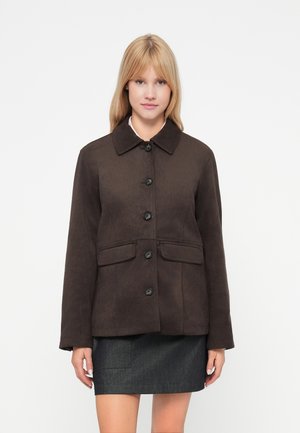 Veste marron texturée avec un col, des boutons à l'avant et deux poches. Portée avec une jupe gris foncé. Design simple et carré.