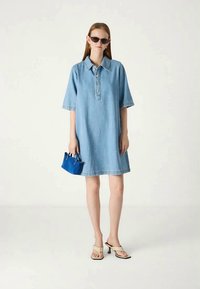 Vestido de camisa de ganga em azul claro, com mangas curtas, gola, fecho de botões na frente e corte reto. Combinado com uma pequena bolsa azul e sandálias.