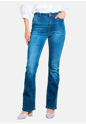 Jeans in denim blu a vita alta con un design a gamba svasata, dotati di cinque tasche e leggermente sbiaditi, abbinati a una blusa bianca e scarpe bianche.