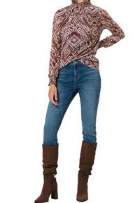 Blouse à manches longues avec un col montant, présentant un motif géométrique multicolore dans des teintes de bourgogne, marron et crème. Associée à un jean bleu et des bottes marron jusqu'au genou.