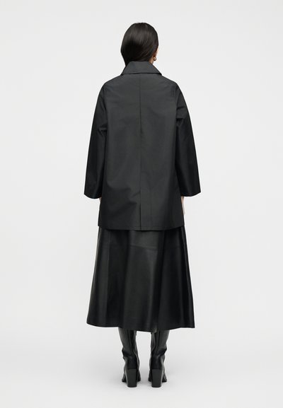 Γυναίκα με μακριά σκούρα μαλλιά που φοράει μαύρο παλτό, μαύρη midi φούστα από δέρμα και μαύρες μπότες με τακούνι, στέκεται μπροστά σε απλό λευκό φόντο.