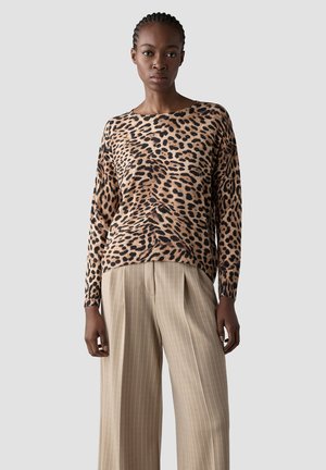 Frau trägt einen langärmeligen Pullover mit Leopardenmuster und beige gestreifte, weit geschnittene Hosen, steht vor einem schlichten hellen Hintergrund.