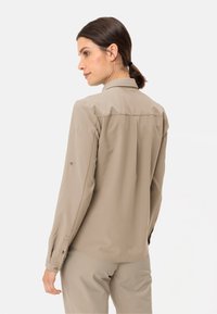 Camicia lunga di colore beige chiaro con colletto, polsini con bottone e orlo curvo. Il tessuto ha una texture liscia con dettagli minimi.