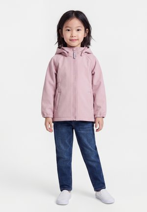 Jong meisje staand, gekleed in een lichtroze gewatteerde jas met capuchon, blauwe spijkerbroek en witte instapschoenen tegen een effen witte achtergrond.