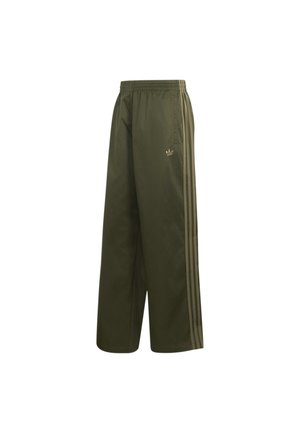 Osoba nosząca luźne oliwkowo-zielone spodnie dresowe Adidas z bocznymi paskami i czarne buty sportowe Adidas z białymi paskami, ręce w kieszeniach.