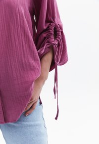 Blusa viola, con texture, maniche gatherate e legacci, indossata su denim azzurro chiaro. Focus sui dettagli delle maniche e sul motivo del tessuto.