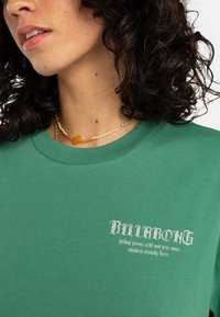 Camiseta de algodón verde con cuello redondo, que presenta un logo y texto impresos en blanco. Textura suave con un corte relajado.