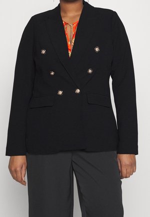 Persona que lleva una blazer negra cruzada con botones dorados sobre una blusa roja con estampado y pantalones gris oscuro sobre un fondo liso.