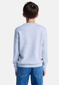 Un bambino con i capelli corti castani indossa una felpa grigia e jeans blu, visto da dietro su uno sfondo bianco.