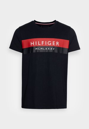 T-shirt en coton marine à manches courtes, avec une large bande horizontale rouge et noire portant les inscriptions "HILFIGER" et "MCMLXXXV" en blanc.
