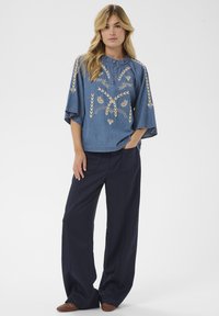 Naine kannab sinist tikandiga laia pluusi, tumeda lai jalgadega pükse ja pruune kootud jalanõusid, seistes üks käsi taskus valge tausta ees.