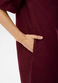 Burgundin väri puuvillavaatekappale, jossa on sivutasku ja lyhyet hihat, esittelee sileän tekstuurin ja muotoillun istuvuuden.