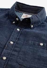 Chemise bleu marine boutonnée avec motif en croisillons blancs, boutons en nacre, poche poitrine et étiquette affichant la marque et un logo de palmier à l'intérieur du col.