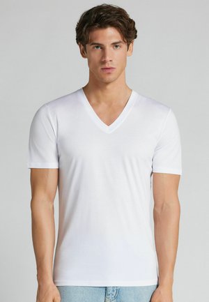 Jonge man met kort krullend haar, die een effen wit V-hals T-shirt met korte mouwen en lichtblauwe spijkerbroek draagt op een grijze achtergrond.