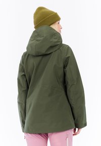 Persoon in een olijfgroene hoodie en mosterdkleurige gebreide beanie, gezien vanaf de achterkant, gecombineerd met lichtroze broek tegen een witte achtergrond.