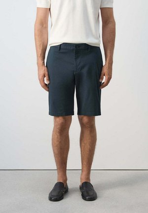 Hombre con pantalones cortos azul oscuro, camisa blanca de manga corta y zapatos sin cordones negros, de pie sobre suelo gris frente a una pared blanca.