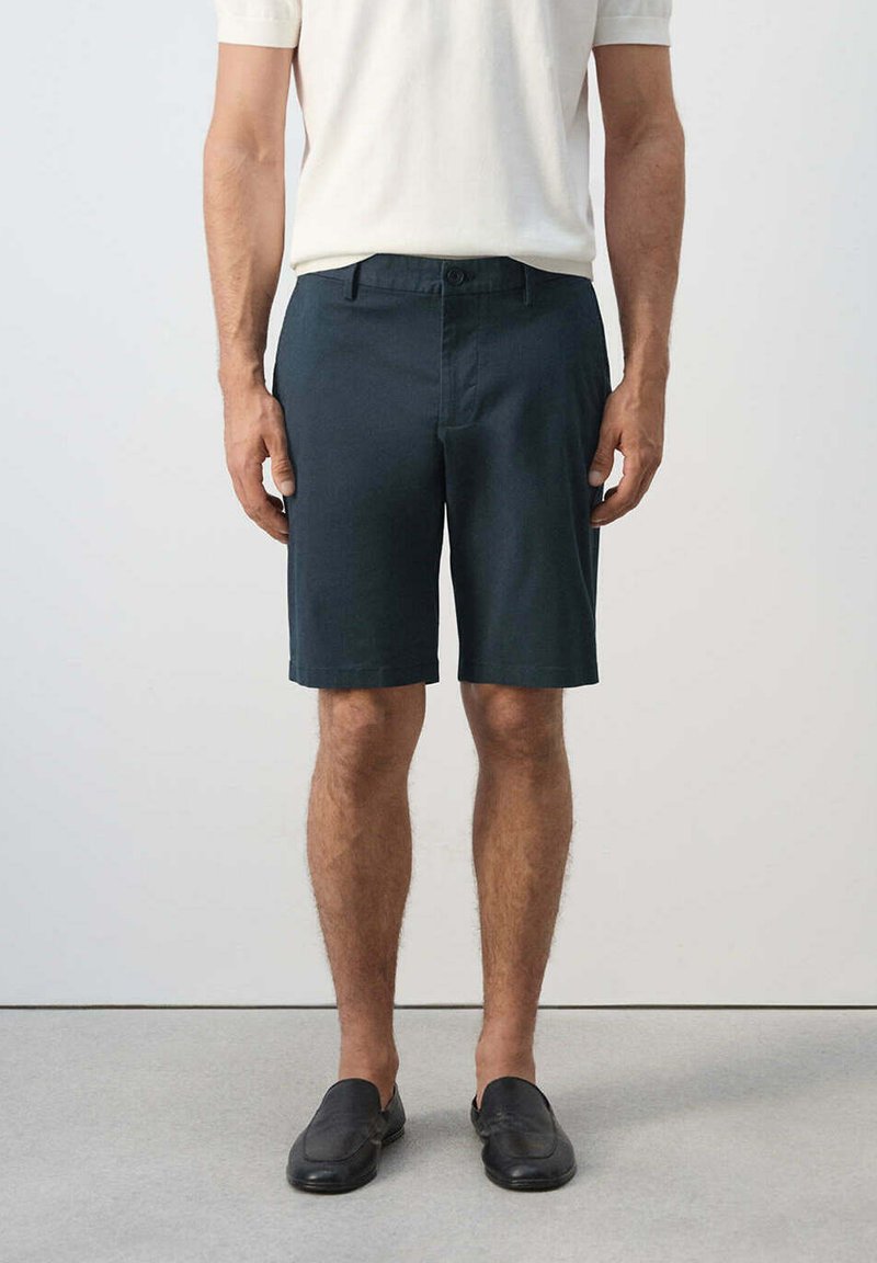 Homme portant un short bleu foncé, une chemise blanche à manches courtes et des chaussures noires sans lacets, debout sur un sol gris contre un mur blanc.