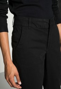 Un pantalon en coton noir présente une texture lisse, une taille plate et des poches latérales. La ceinture est dotée de passants de ceinture et offre une coupe propre et ajustée.