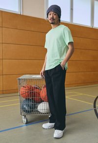 Mintgrøn kortærmet skjorte, sorte vide bukser, hvide sneakers. I baggrunden en vogn fyldt med forskellige basketballer og en basketballbane.