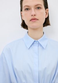 Camicia button-up azzurro chiaro con colletto classico, texture liscia e maniche lunghe. Presenta una chiusura a bottoni e una vestibilità rilassata.