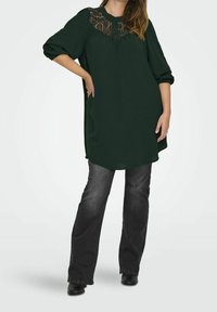 Mörkgrön tunika med spetsdetaljer vid halsen, med en knappslå, tillsammans med svarta bootcut-jeans och anklarstövlar.