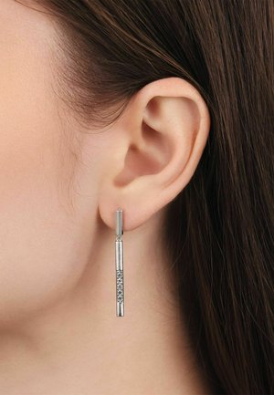 Primer plano de la oreja de una mujer que lleva un pendiente largo y delgado de plata con una sección central texturizada y brillante. Se ve cabello castaño.