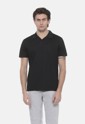 Uomo con capelli castani indossa una polo nera a maniche corte e jeans grigio chiaro, in piedi davanti a uno sfondo bianco uniforme.