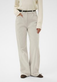 Sieviete, kas valkā gaiši bežīgas platās bikses, iekļautu krēmkrāsas džemperī ar garām piedurknēm, melnu jostu un brūnām plakanu apavu, stāvot ar vienu roku kabatā.