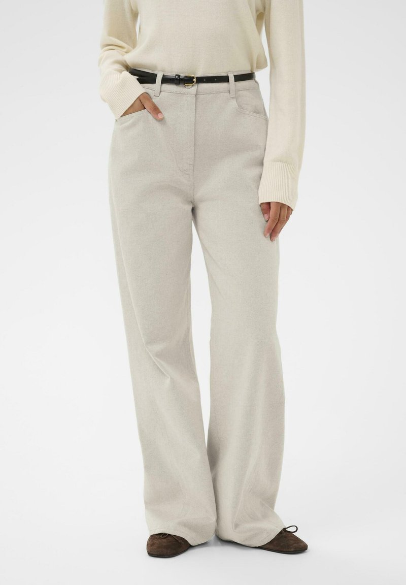 Sieviete, kas valkā gaiši bežīgas platās bikses, iekļautu krēmkrāsas džemperī ar garām piedurknēm, melnu jostu un brūnām plakanu apavu, stāvot ar vienu roku kabatā.