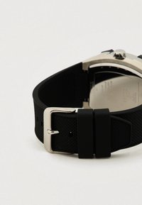 Bracelet de montre en caoutchouc noir avec une surface texturée, boucle en métal argenté et fond de boîtier en acier inoxydable visible de l'autre côté.