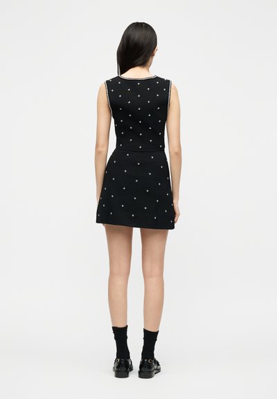 Γυναίκα που φοράει μαύρο φόρεμα χωρίς μανίκια με λευκά πουά και διακόσμηση, συνδυασμένο με μαύρες κάλτσες και γυαλιστερά μαύρα μοκασίνια, όρθια με την πλάτη στραμμένη προς το μέρος.