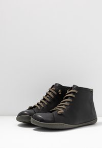 Černé kožené kotníkové boty s šedými podrážkami, vybavené hnědými tkaničkami a jednoduchým, zaobleným designem. Texturovaná kůže na bocích dodává detail.