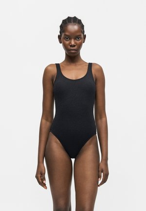 Femme aux cheveux tressés portant un maillot de bain une-pièce noir texturé, debout droite devant un fond clair uni.