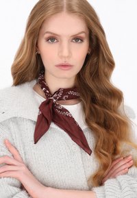 Pălăria de tip bandană, de culoare maro, are un model paisley alb. Este purtată legată în jurul gâtului peste un cardigan tricotat gri, cu un design texturat.
