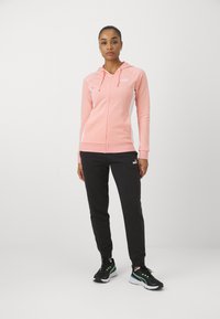 Roza zip-up dukserica s kapuljačom na vezanje, bijele bočne panele i crne trenerke. Model nosi crne tenisice s zelenim akcentima.