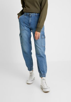 Osoba ubrana w niebieskie dżinsowe spodnie cargo typu jogger z elastycznymi ściągaczami, białe wysokie trampki oraz oliwkową, długorękawową bluzę, stojąca.