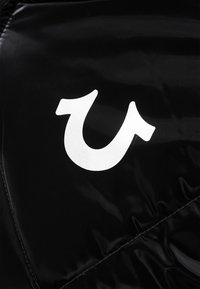 Logo blanc stylisé en forme de fer à cheval sur un tissu noir brillant, probablement une partie d'un vêtement ou d'un accessoire.