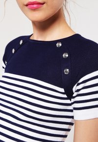 Top de punto a rayas en azul marino y blanco con mangas cortas, que presenta botones decorativos plateados en el hombro y una textura suave y ajustada.