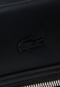 Μαύρη επιφάνεια από υφή δέρματος με ένα μικρό μαύρο έμβλημα κροκοδείλου πάνω από μια ασημένια λεπτομέρεια φερμουάρ.