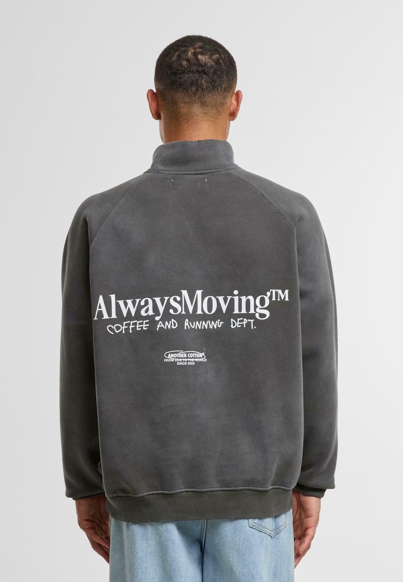 Felpa grigia con colletto alto, con grande testo bianco sul retro: "AlwaysMoving™ COFFEE AND RUNNING DEPT." Design semplice e privo di decorazioni.