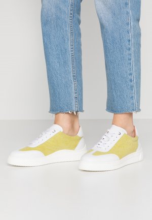 Person wearing light blue jeans and white sneakers with yellow suede panels standing on a white floor.