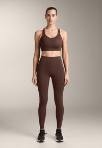 Nő áll szemben a nézővel, barna sportmelltartót, magas derekú leggingset, fekete zoknit és sötét tornacipőt visel egy egyszerű háttér előtt.