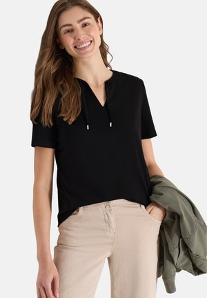 Donna sorridente con capelli lunghi che indossa un top nero a maniche corte e pantaloni beige, tenendo una giacca verde oliva nella mano sinistra.