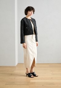 Giacca in tweed nera cropped con due tasche frontali, abito senza maniche crema con spacco frontale e mule nere open-toe con tacco basso.