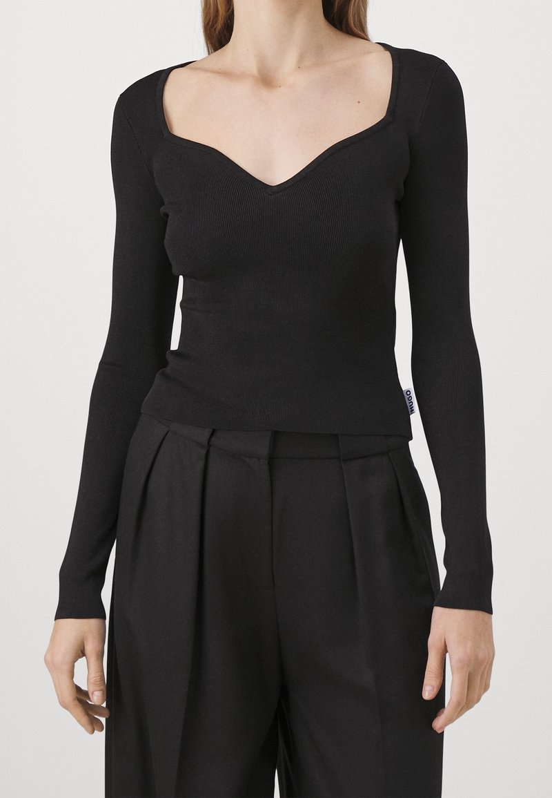 Femme portant un haut noir ajusté à manches longues avec un décolleté en cœur et un pantalon plissé noir taille haute.