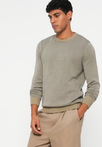 Sweater de malha beige com padrão, mangas longas, decote redondo e punhos canelados. Usado com calças castanho claro. A textura é suave e entrelaçada.