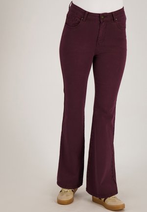 Donkerpaarse, hoge taille broek met uitlopende pijpen, gedragen met beige en bruine sneakers op een neutrale achtergrond.