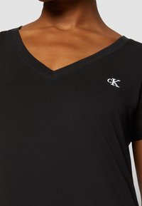 Černé bavlněné tričko se V-výstřihem, krátkými rukávy a malým bílým logem "cK" na levé části hrudi.